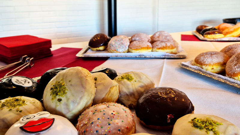 Auf dem Foto sind verschiedene Berliner (Krapfen, Pfannkuchen) mit unterschiedlichen Glasuren und Füllungen zu sehen, die auf Tellern auf einem gedeckten Tisch arrangiert sind. Einige Berliner haben Schokoladenüberzug, andere sind mit Zuckerguss oder Puderzucker bestäubt, und einige sind mit bunten Streuseln oder Pistazien dekoriert. Im Hintergrund gibt es einen runden Stehtisch mit mehreren Flaschen eines Getränks. Rote Servietten sind ordentlich gestapelt. Die Umgebung wirkt modern mit einer weißen Backsteinwand im Hintergrund.