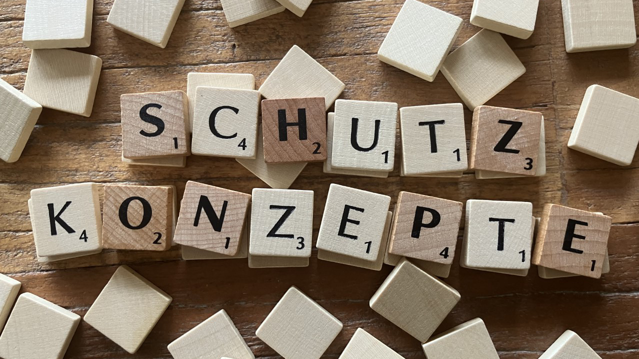 Auf dem Foto sind Scrabble-ähnliche Holzbuchstabensteine zu sehen, die auf einem hölzernen Untergrund verteilt sind. Einige der Steine sind zu den Wörtern „SCHUTZ KONZEPTE“ zusammengesetzt, während andere verstreut herumliegen.