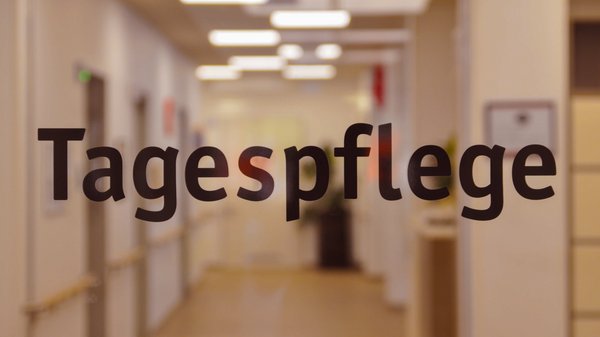 Zu sehen ist ein unscharf fotografierter, hell beleuchteter Flur einer Einrichtung. Im Vordergrund steht auf einer Glastür oder Glasscheibe groß der Schriftzug „Tagespflege“. Im Hintergrund erkennt man unter anderem Deckenleuchten, Türen, Handläufe und einen Flurbereich.
