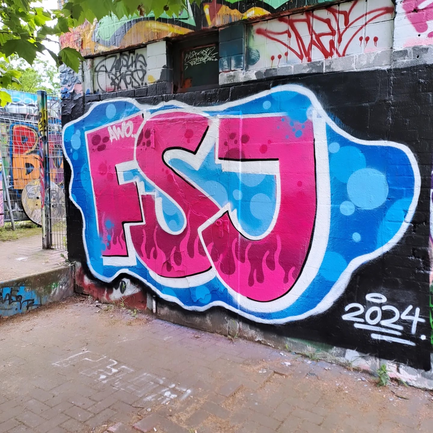 Das Foto zeigt ein Graffiti auf einer Wand. Das Graffiti besteht aus stilisierten Buchstaben, die “FSJ” in großen, blasenartigen Zeichen mit einem 3D-Effekt buchstabieren. Die Buchstaben sind hauptsächlich in Blau- und Pinktönen gemalt, mit weißen Highlights, die den Eindruck von Glanz oder Reflexion erwecken. Unter den Buchstaben befindet sich eine kleinere Inschrift, die “2024” lautet. Die Hintergrundwand ist mit verschiedenen anderen Graffiti-Tags und Markierungen bedeckt, was eine vielschichtige Textur von Straßenkunst erzeugt.