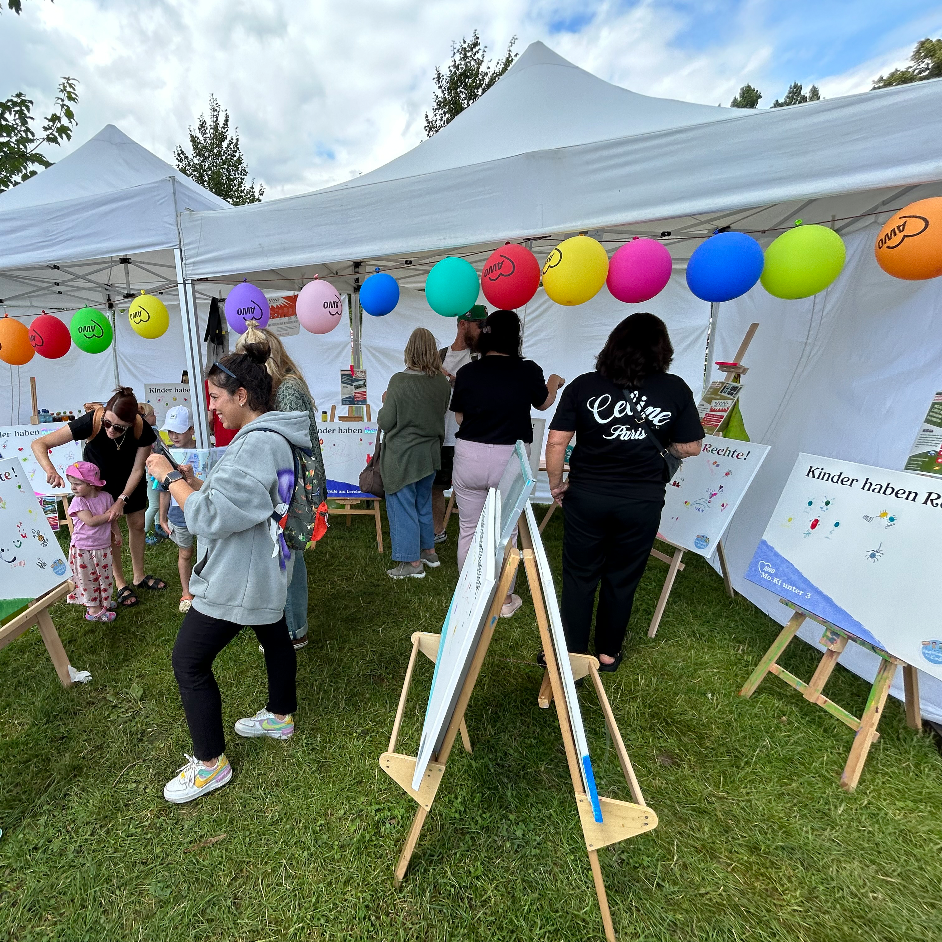 Das Bild zeigt eine Outdoor-Veranstaltung mit mehreren Personen, darunter Kinder und Erwachsene, die an Aktivitäten teilnehmen. Es gibt weiße Zelte mit bunten Luftballons daran. In den Zelten stehen Staffeleien mit Papieren, auf denen es so aussieht, als könnten die Menschen malen oder zeichnen.