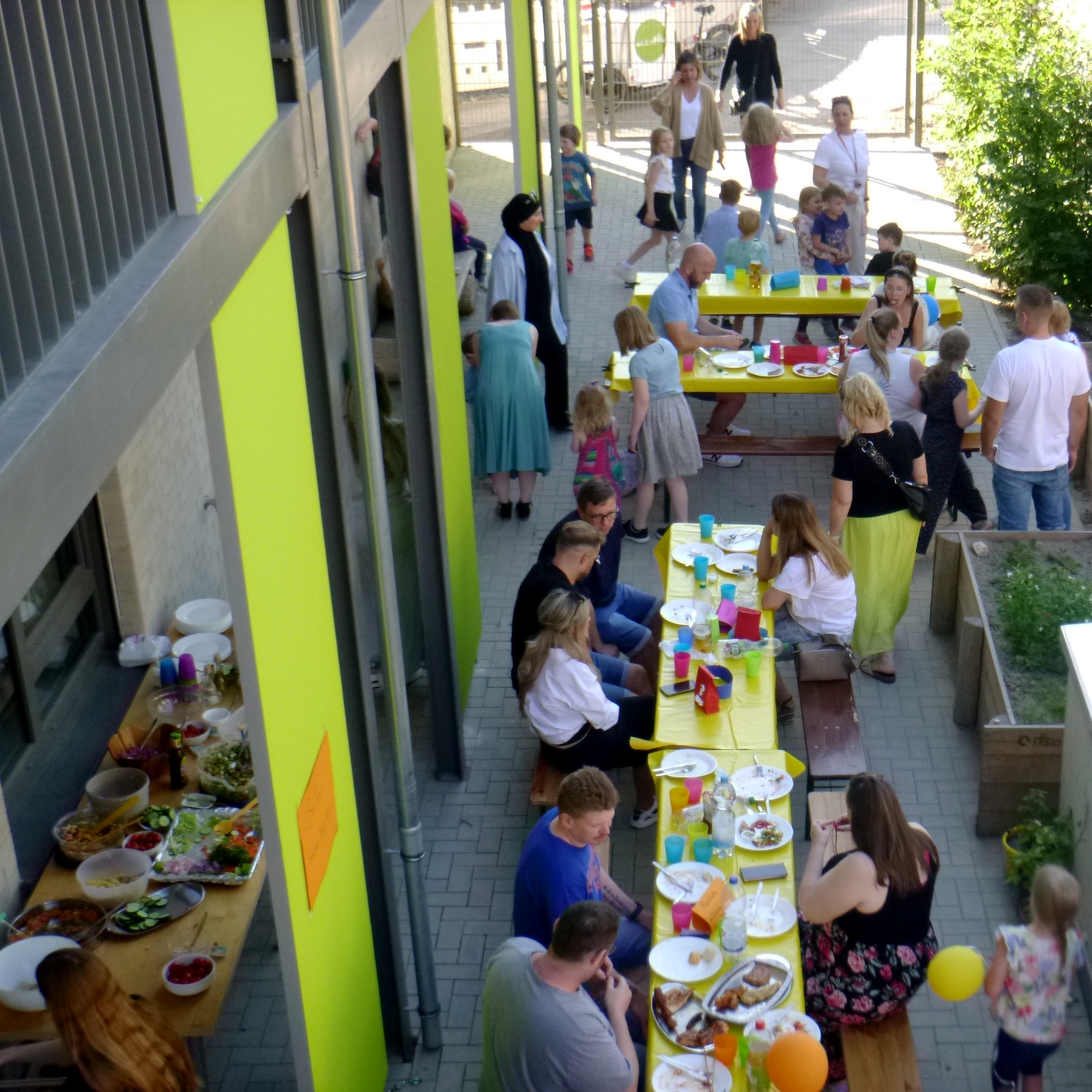 Das Foto zeigt eine Party. Menschen unterschiedlichen Alters sitzen an Tischen. Die Tische sind mit bunten Dekorationen und Essen gedeckt. Die Szenerie wirkt sonnig und spielt sich vor einem Gebäude mit grünen und gelben Wänden ab. 