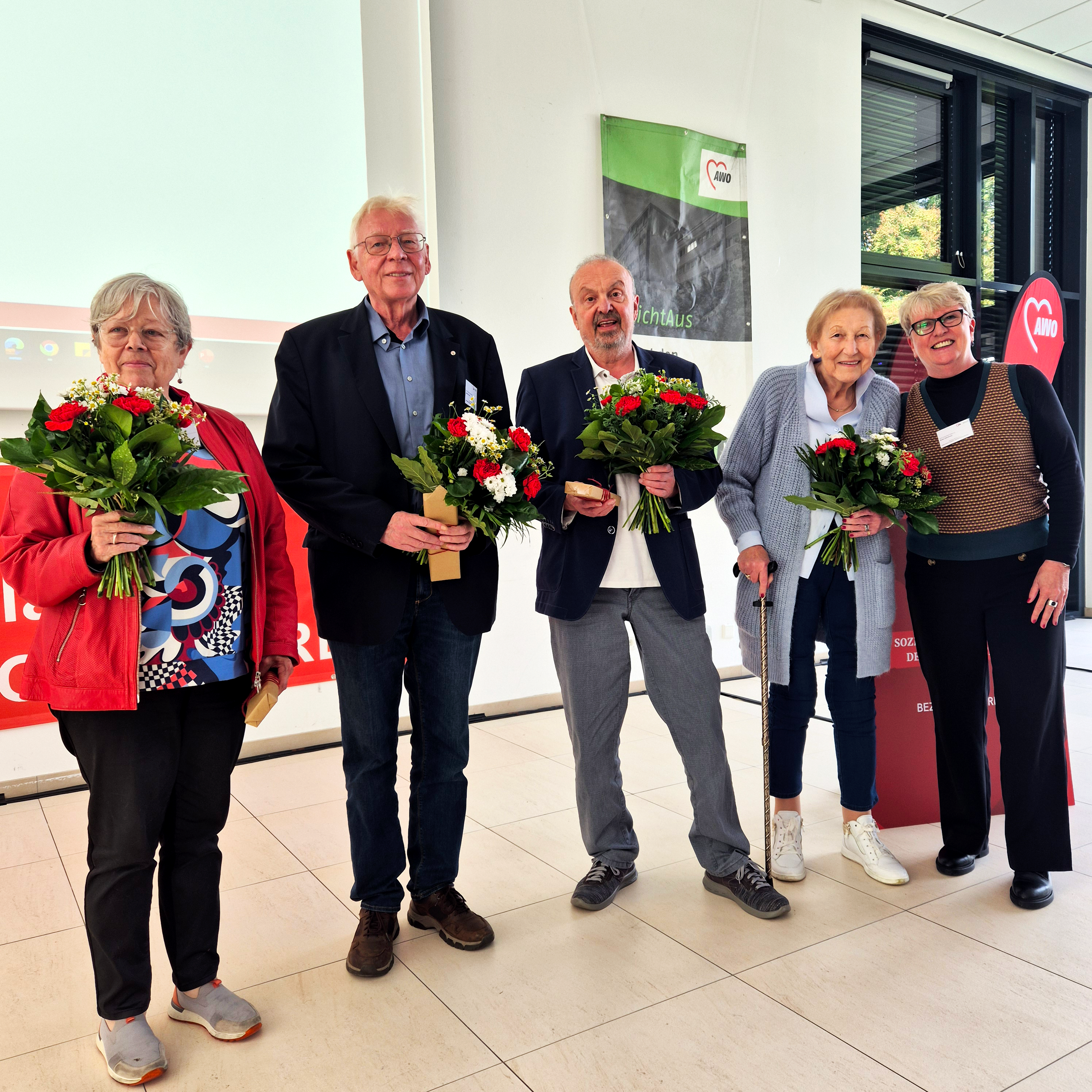 Das Foto zeigt eine Gruppe von sechs Personen, die in einem Innenraum stehen und jeweils einen Blumenstrauß halten. 