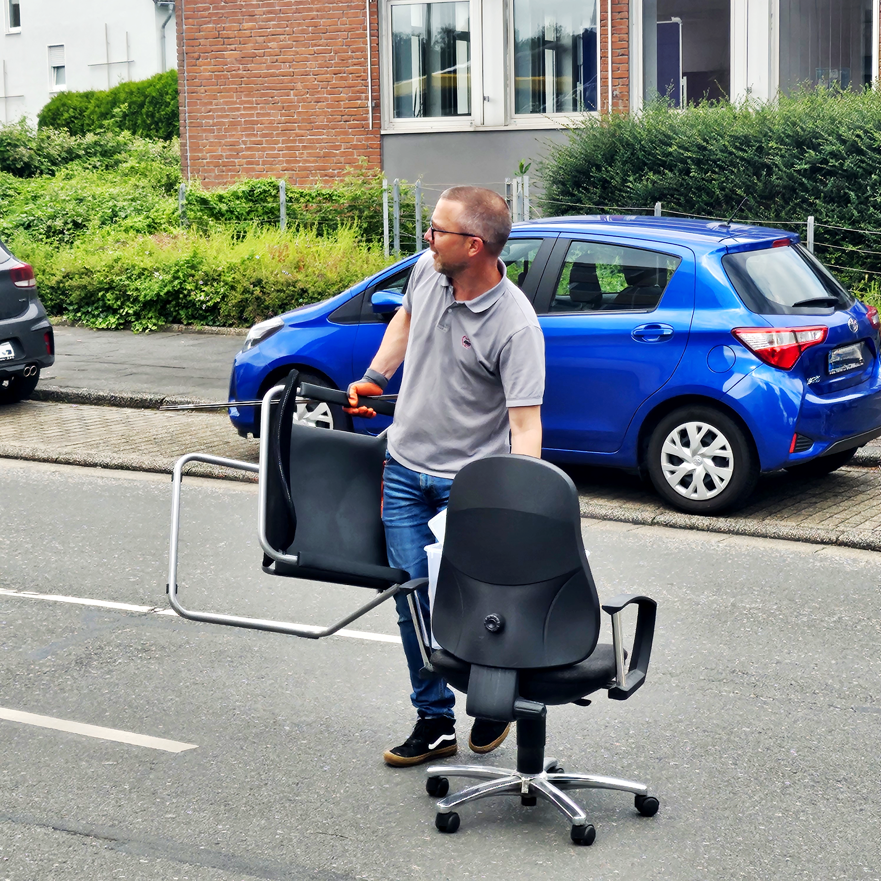 Das Foto zeigt eine Person, die zwei Bürostühle über eine Straße schiebt. Die Person trägt ein graues Hemd und Jeans. Hinter der Person ist ein blauer geparkter Wagen zu sehen, und im Hintergrund sind Gebäude mit Grünflächen erkennbar.
