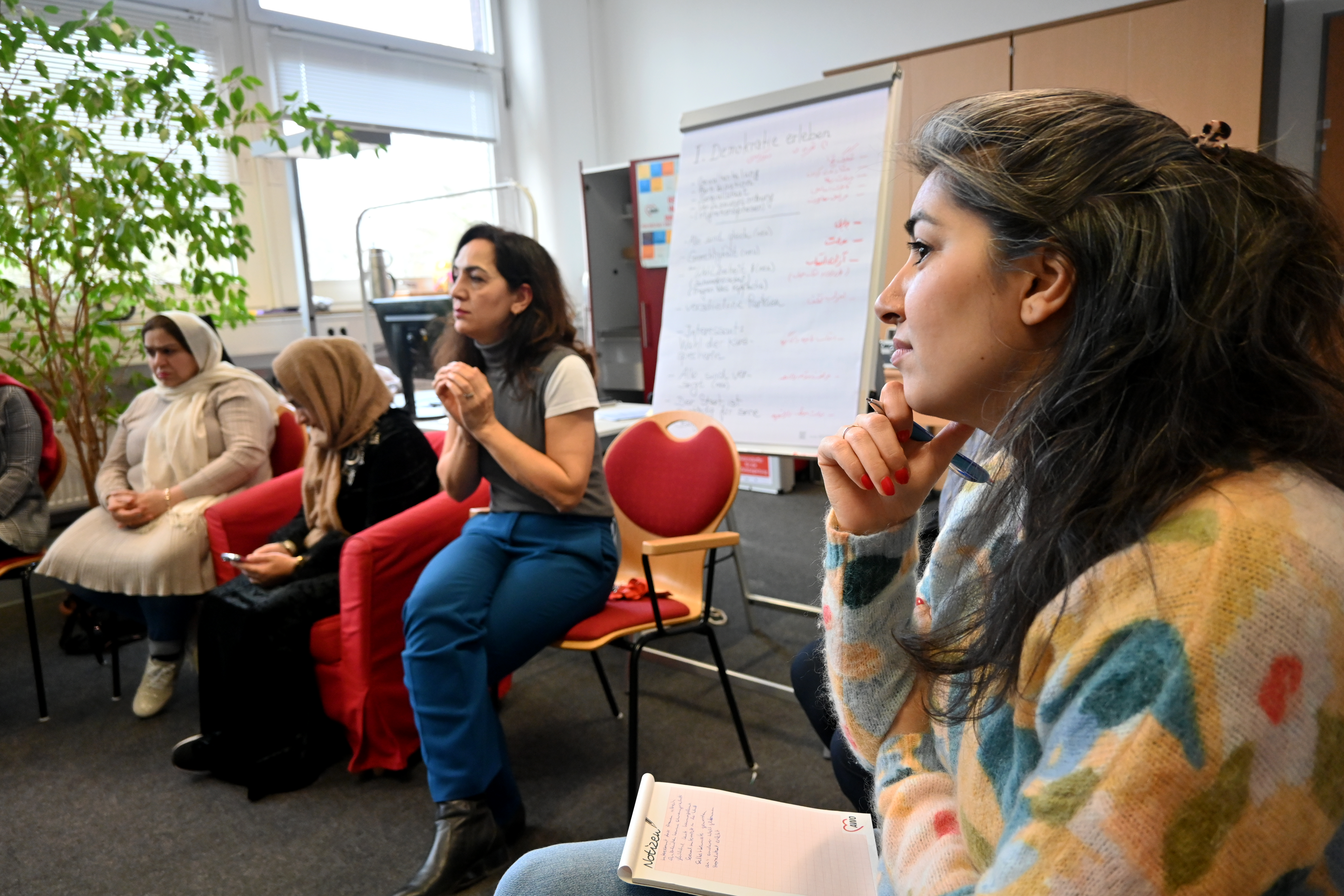 Das Foto zeigt eine Gruppe von Frauen, die in einem Raum zusammensitzen und an einer Diskussion oder einem Workshop teilnehmen. Im Vordergrund ist eine Frau mit langen, dunklen Haaren zu sehen, die konzentriert zuhört und Notizen auf einem Block macht. Im Hintergrund sind weitere Teilnehmerinnen zu erkennen, darunter eine Frau, die aktiv spricht und gestikuliert. Es gibt eine Flipchart mit handgeschriebenem Text.