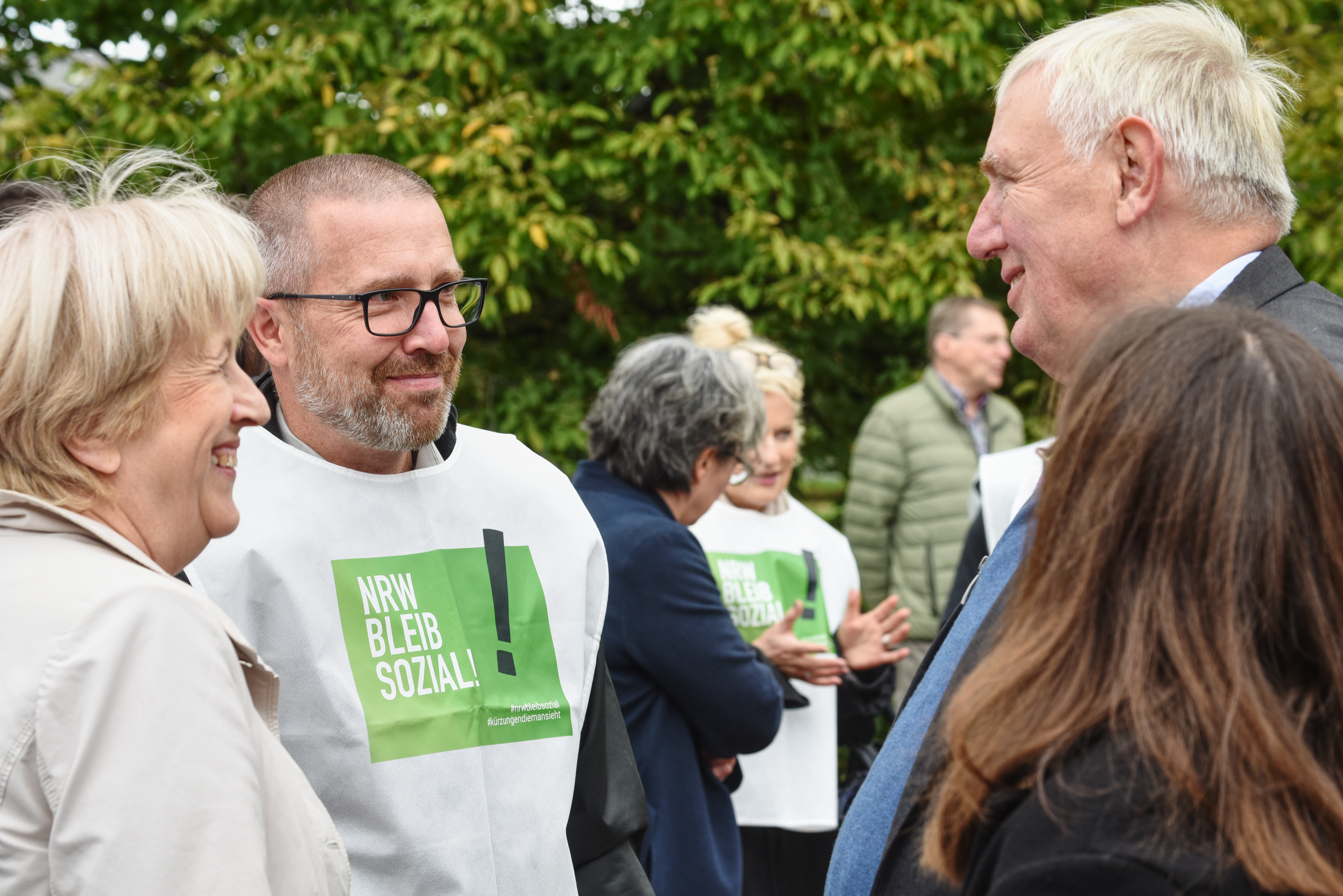 Auf dem Foto ist eine Gruppe von Personen im Freien zu sehen. Eine Person im Vordergrund trägt eine weiße Weste mit grünem Text, auf dem “NRW BLEIB SOZIAL” steht. 