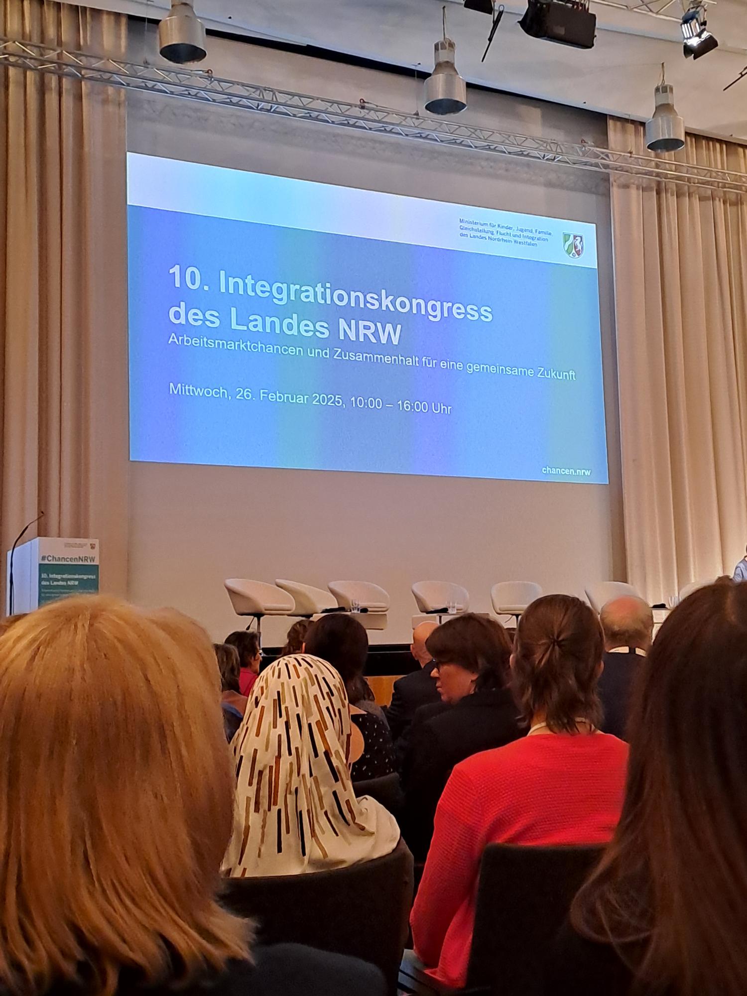 Das Foto zeigt eine Veranstaltung, den 10. Integrationskongress des Landes NRW. Es handelt sich um eine Konferenz mit dem Titel "Arbeitsmarktchancen und Zusammenhalt für eine gemeinsame Zukunft", die am 26. Februar 2025 von 10:00 bis 16:00 Uhr stattfindet. Auf der Bühne stehen fünf leere Stühle für eine Podiumsdiskussion, und das Publikum sitzt bereits im Saal.