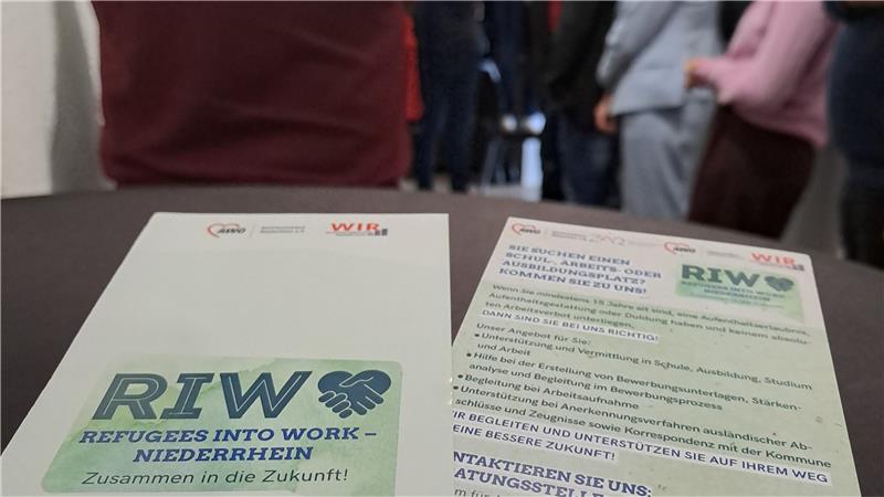 Das Foto zeigt eine Veranstaltung oder ein Treffen mit mehreren Personen, die in einer Reihe oder Gruppe stehen und miteinander sprechen. Im Vordergrund liegen auf einem Tisch zwei Flyer oder Broschüren mit dem Titel "RIW – Refugees Into Work – Niederrhein". Die Unterzeile lautet "Zusammen in die Zukunft!", was auf ein Integrations- und Arbeitsmarktprojekt für Geflüchtete hinweist. Die Flyer enthalten Informationen zur Unterstützung von Geflüchteten beim Einstieg in den Arbeitsmarkt, etwa durch Beratung, Bewerbungshilfen und Qualifizierungsmaßnahmen. Im Hintergrund sind Personen zu sehen, die scheinbar an einer Diskussion oder einem Networking-Event teilnehmen.