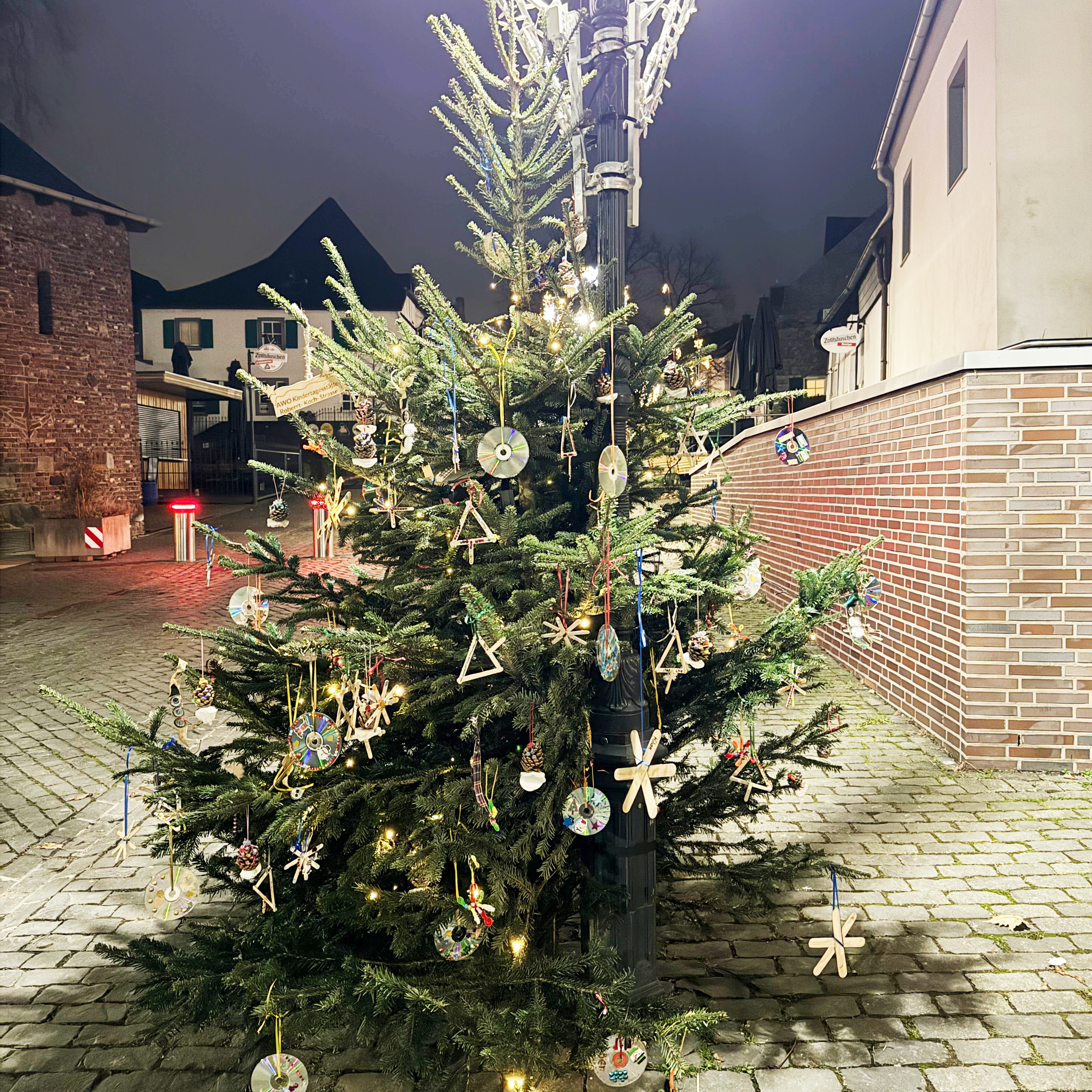 Das Foto zeigt einen geschmückten Weihnachtsbaum, der sich im Freien auf einem gepflasterten Platz befindet. Der Baum ist mit verschiedenen Dekorationen behängt, darunter CDs, Holzfiguren und andere handgefertigte Ornamente. Im Hintergrund sind Gebäude und eine Straßenlaterne zu erkennen, die den Bereich beleuchtet.